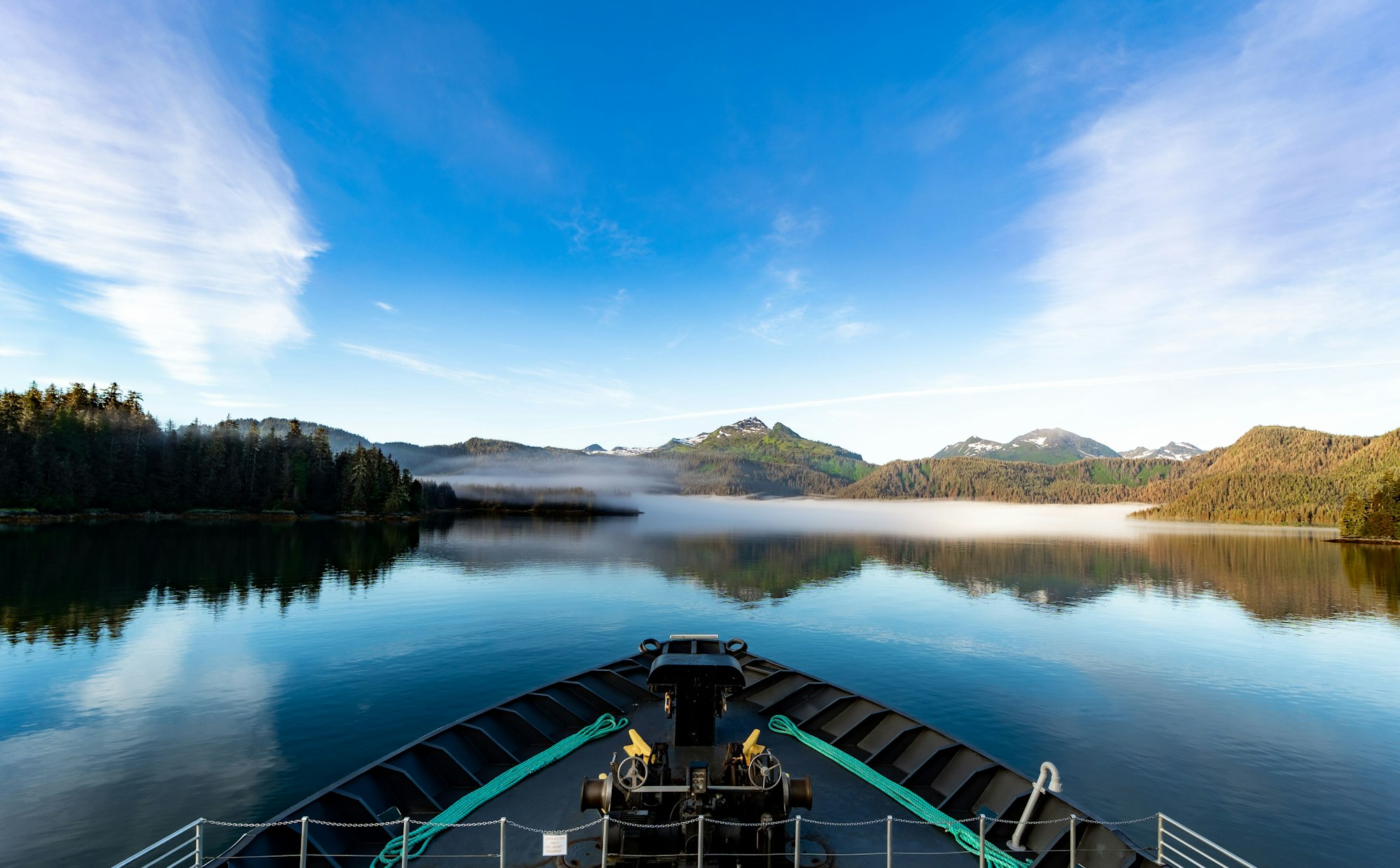 Boat view of landscape with hills and trees reflected in lake water under blue sky with white clouds